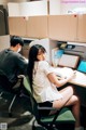 A woman sitting at a desk in an office with a man in the background.